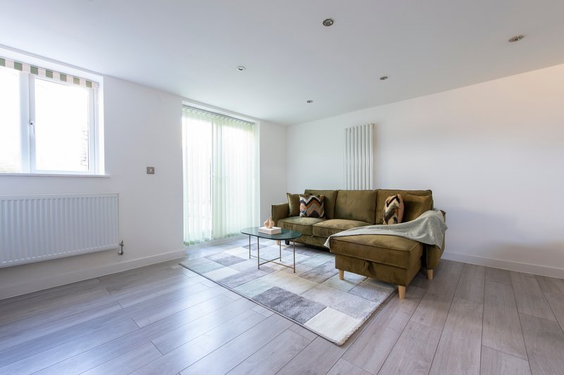 Bright living room with olive-green corner sofa, glass coffee table, wooden flooring, and large windows