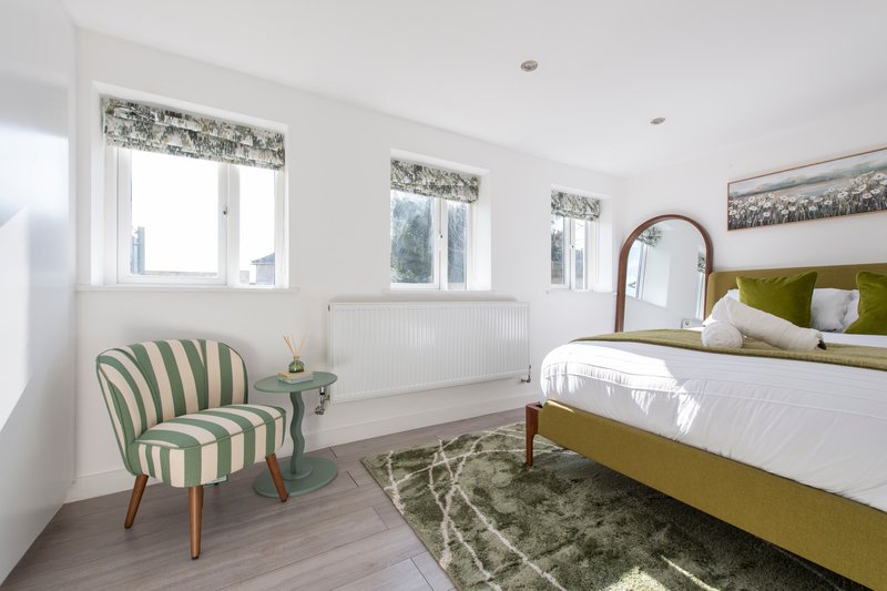 Bright bedroom with olive-green upholstered bed, striped armchair, wooden flooring and windows