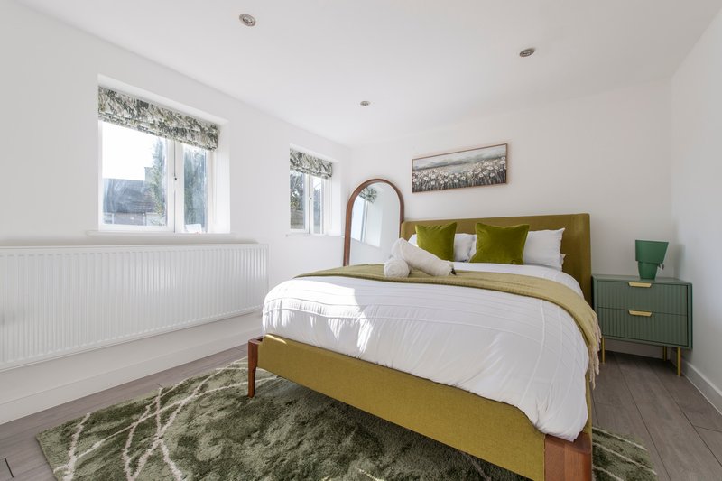 Bright bedroom with olive-green upholstered bed, white bedding, wooden nightstand, and windows with patterned curtains