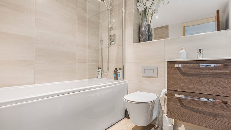 Modern bathroom with white tile walls, bathtub with glass screen, toilet, and wooden vanity