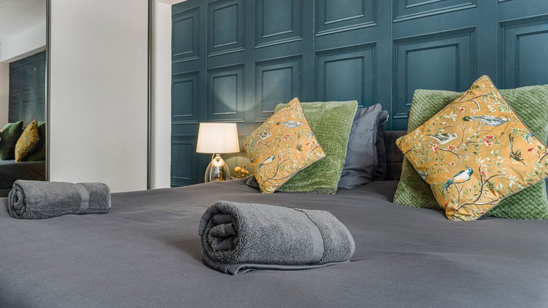 Bedroom with grey bed, decorative yellow and green cushions, rolled towels, and blue panelled headboard