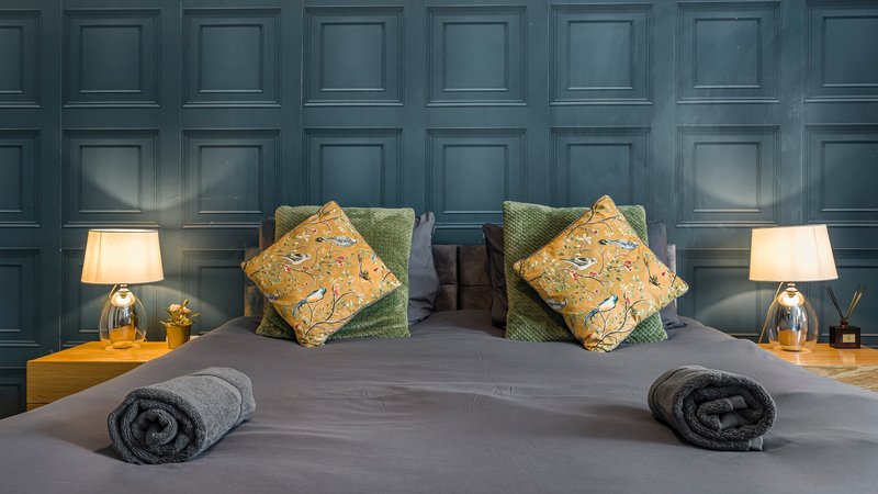Modern bedroom with dark blue panelled headboard, grey bed, patterned cushions and bedside lamps