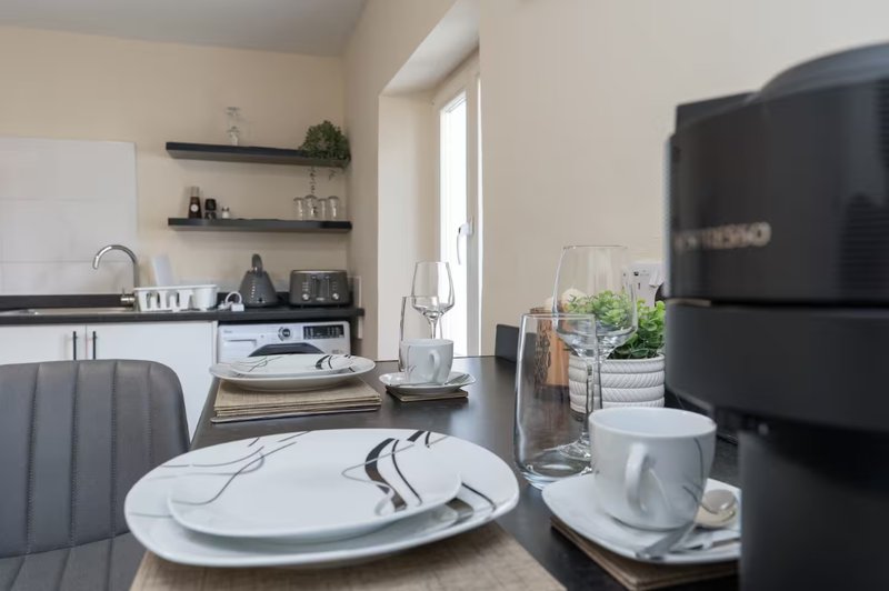 Modern kitchen-diner with white dining table, black appliances, open shelving, and natural light from door