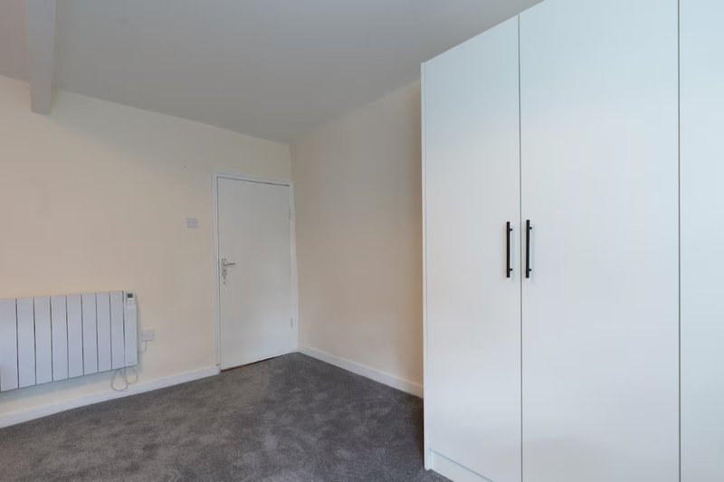 Empty bedroom with white wardrobe, radiator, door and dark carpet flooring