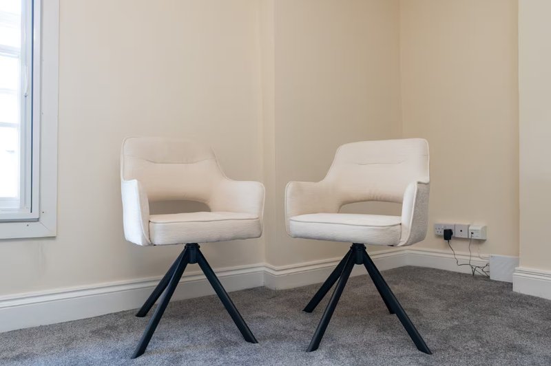 Two cream upholstered chairs with black tripod legs on grey carpet in bright room