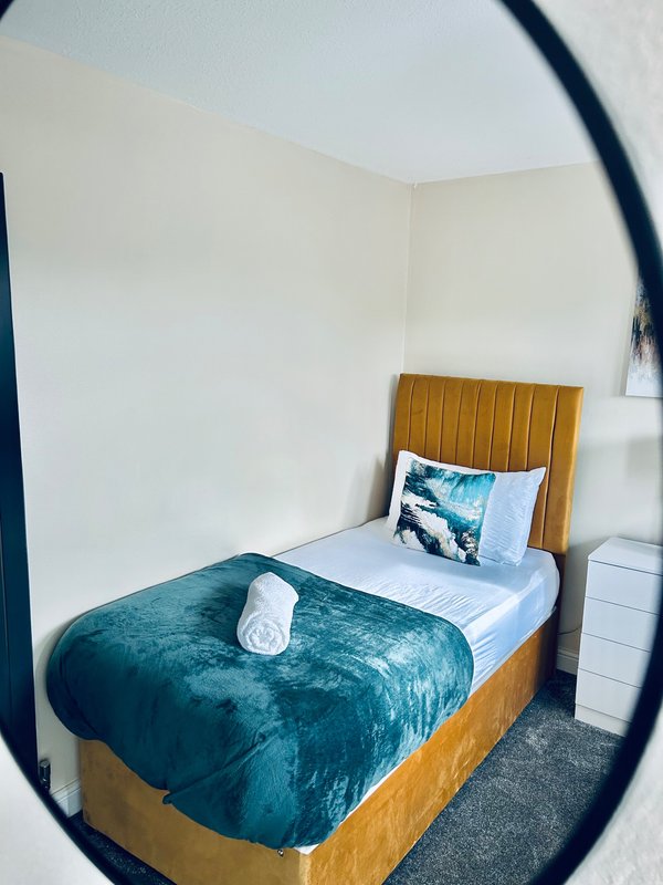 Compact bedroom with single wooden bed, teal velvet bedding, white pillows and rolled towel