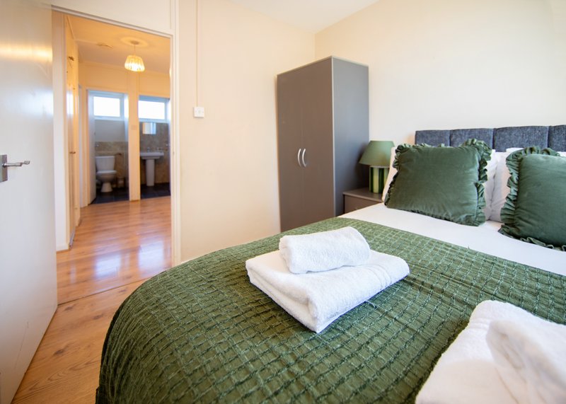 Bright bedroom with double bed, green bedding, white pillows, wooden floor and open doorway