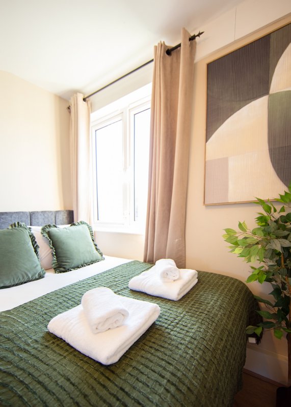Bedroom with green double bed, white towels, green cushions, beige curtains and potted plant