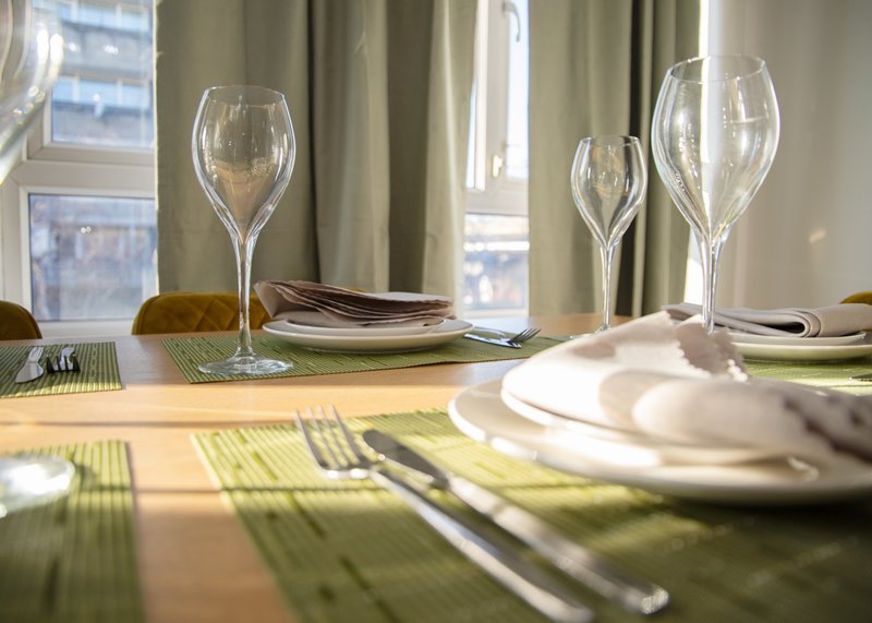 Dining table set with glasses, plates, cutlery and green placemats near windows