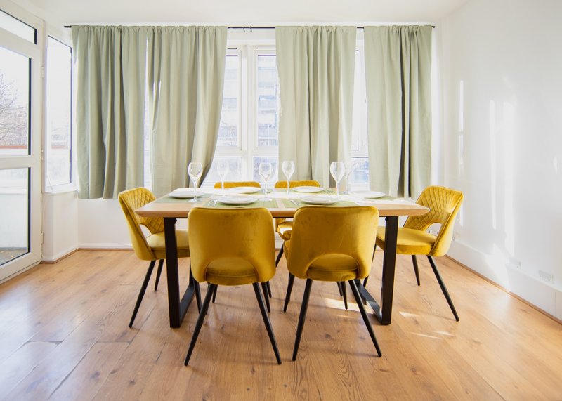 Modern dining room with round wooden table, yellow velvet chairs, large windows with cream curtains