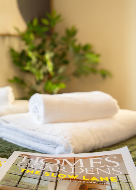 Rolled white towels stacked on bed with plant and magazine in background