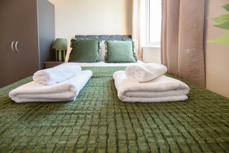 Bedroom with green bedspread, folded white towels and linens on bed, window with curtains