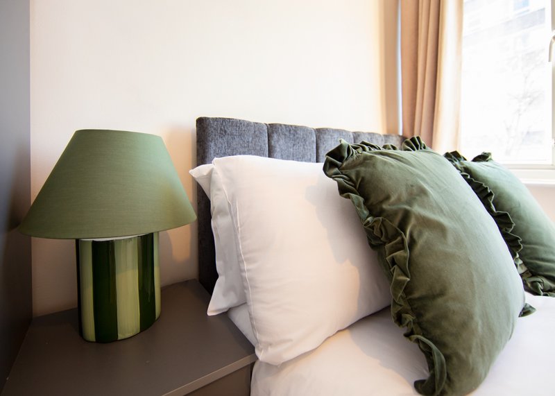 Bedroom with grey upholstered headboard, white pillows, green cushions, and bedside lamp
