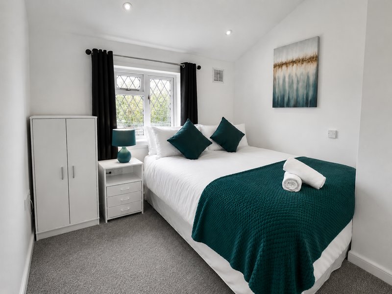 Bright bedroom with double bed, teal bedding, white wardrobe and window with black curtains