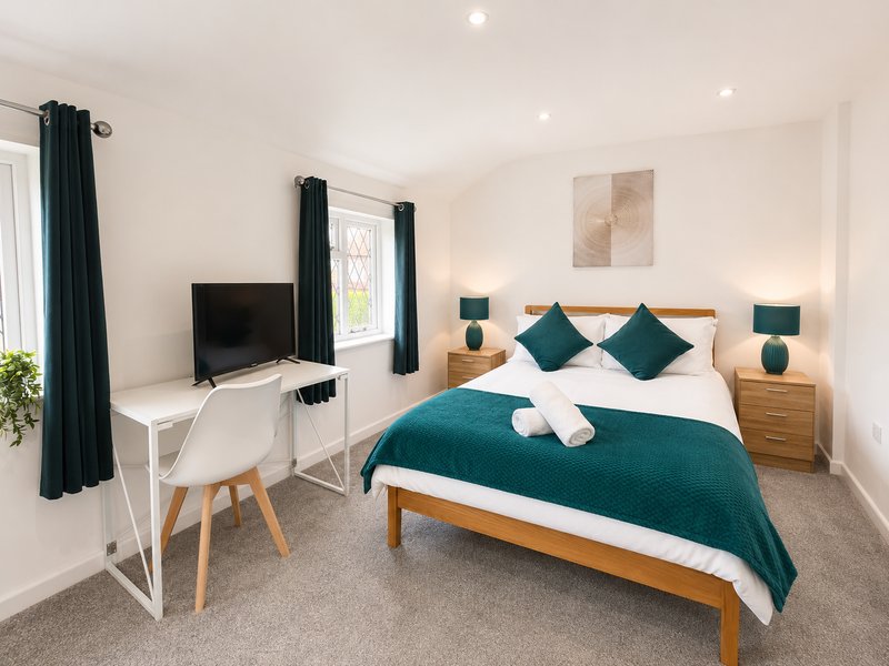 Modern bedroom with double bed, teal bedding, wooden frame, desk with TV, and armchair