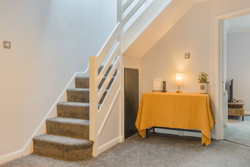 Hallway with carpeted staircase, yellow-draped table, lamp, and doorway to bedroom