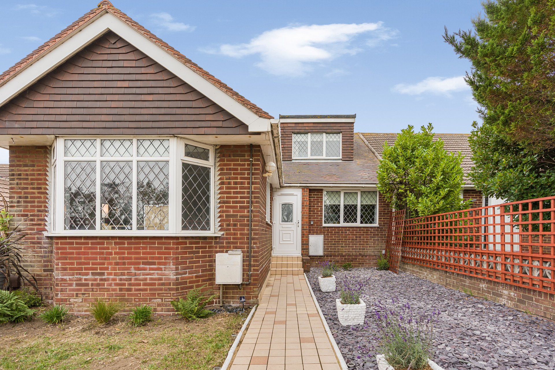 Brick bungalow exterior with lattice windows, gravel driveway, and red fence