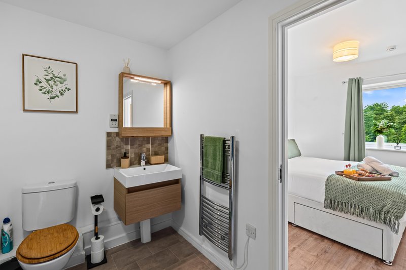 Compact bathroom with wooden vanity, sink, toilet and mirror, adjoining bright bedroom with bed