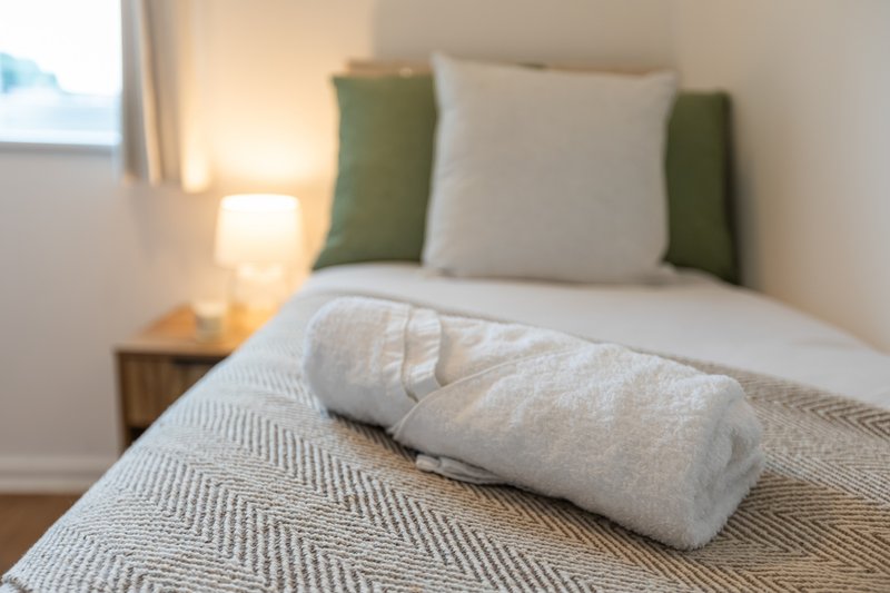 Bedroom with made bed, green and white pillows, rolled towel, bedside lamp and window