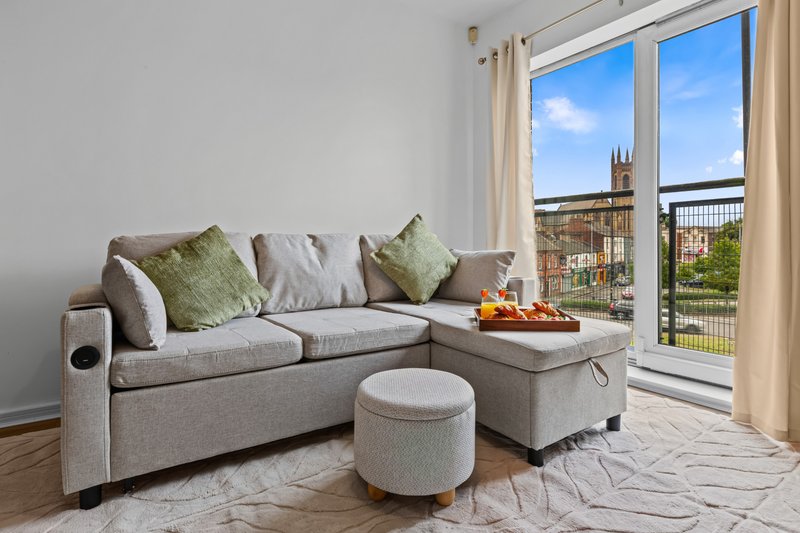Modern living room with gray corner sofa, cushions, ottoman, and balcony doors overlooking cityscape
