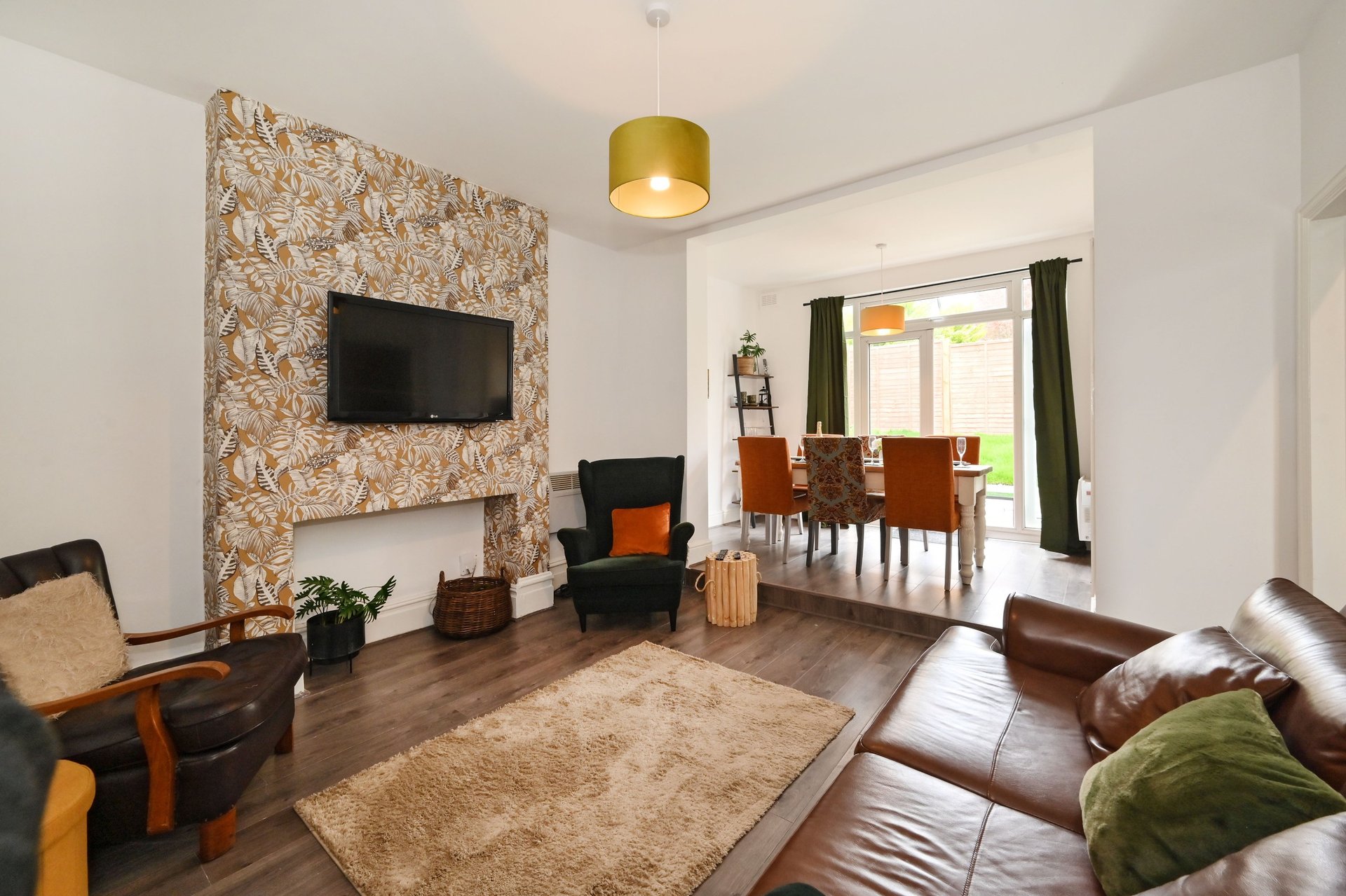 Open-plan living room with feature wallpaper, mounted TV, brown leather sofa and dining area visible through doorway