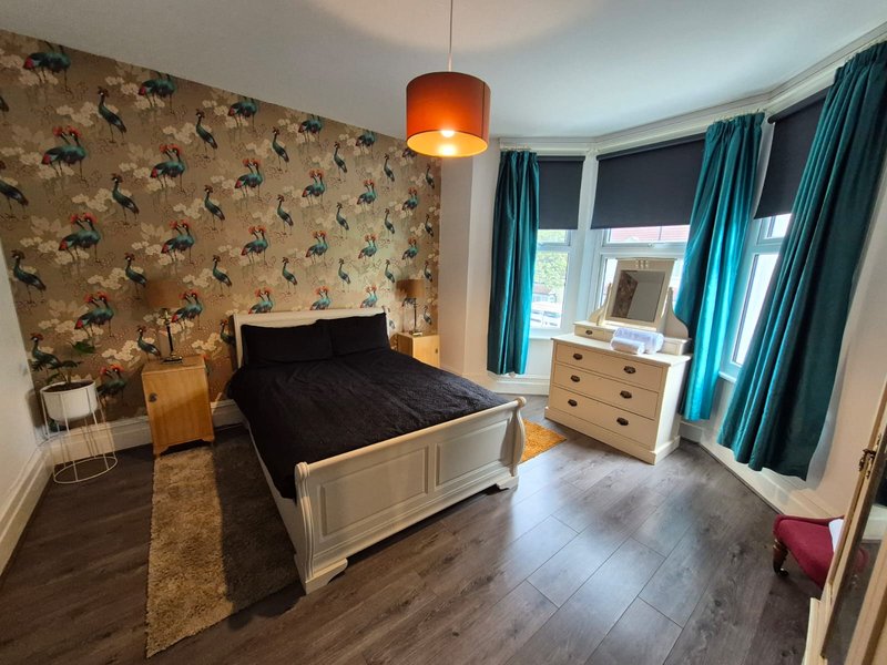Spacious bedroom with double bed, wooden dresser, patterned wallpaper and turquoise curtains