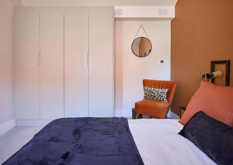 Bedroom with double bed, white wardrobe, orange accent chair and round mirror on wall