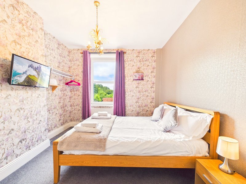 Bright bedroom with wooden bed, white bedding, floral wallpaper, and purple curtains framing window.