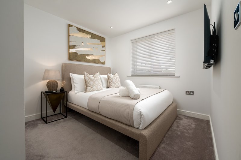 Modern bedroom with neutral upholstered bed, white bedding, decorative pillows, wall art, and mounted television.