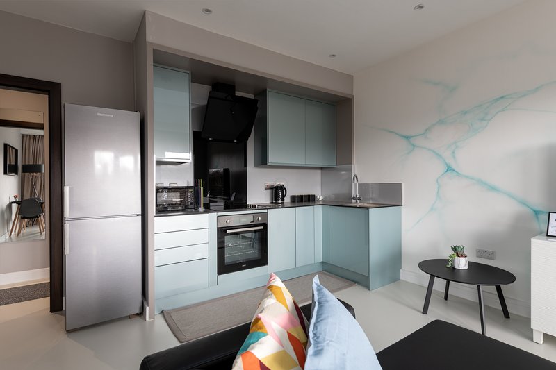 Modern open-plan kitchen and living area with light blue cabinetry, stainless steel appliances, and black sofa with cushions