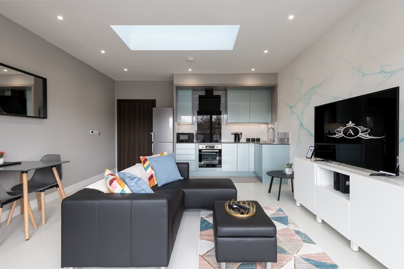 Modern open-plan living room with dark sofa, kitchen area, TV mounted on wall, skylight