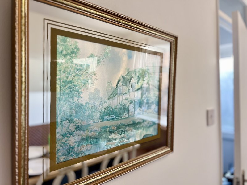 Framed landscape artwork on cream wall depicting countryside house with garden and trees