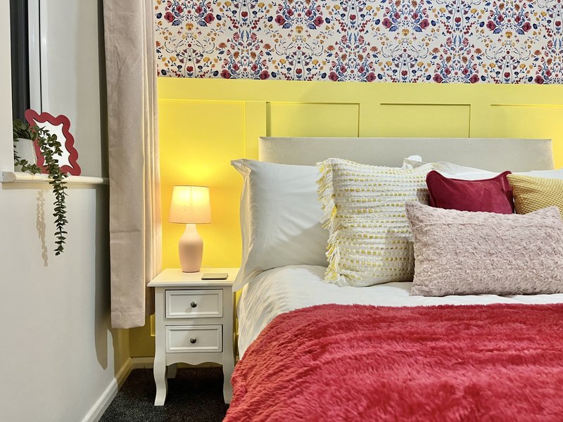 Bedroom with yellow headboard, red bedding, patterned wallpaper, and bedside lamp