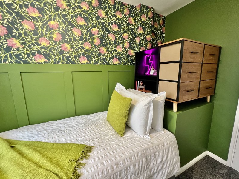Bright green bedroom with double bed, patterned wallpaper, wooden dresser and neon sign