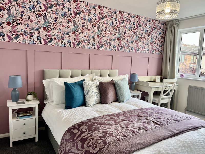 Modern bedroom with pink headboard, patterned wallpaper, double bed with colorful pillows and desk area