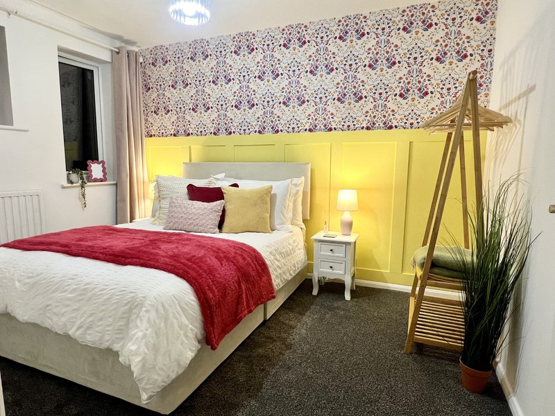 Bedroom with double bed, yellow headboard, patterned wallpaper, potted plant and modern lighting