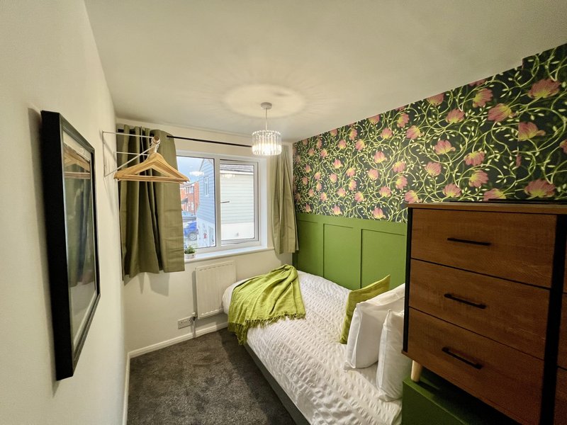 Compact bedroom with single bed, green headboard, wooden dresser and floral wallpaper