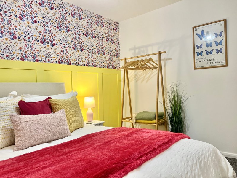Bedroom with yellow headboard, red bedding, patterned wallpaper accent wall, and wooden clothing rack