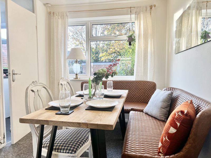 Bright dining area with wooden table, bench seating, cushions and large windows overlooking garden