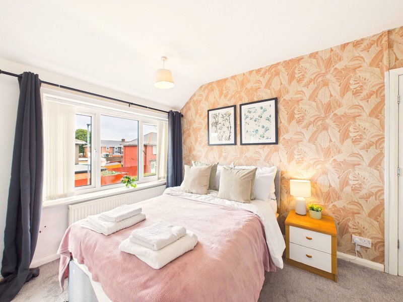 Bedroom with double bed, pink duvet, patterned wallpaper, wooden nightstand and window view