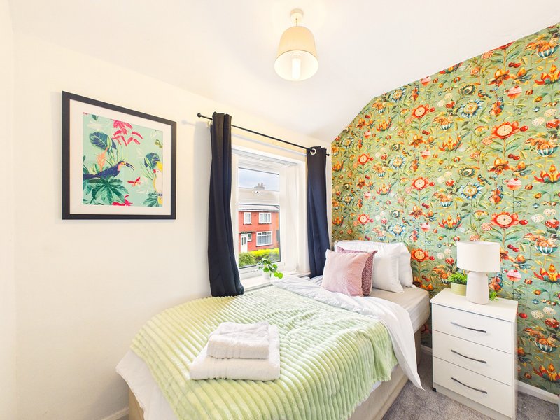Small bedroom with double bed, colorful patterned wallpaper, framed artwork, and bedside table