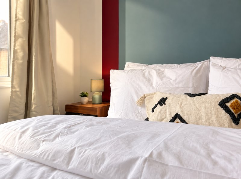 Bedroom with white bedding, decorative cushion, wooden nightstand and window with curtains