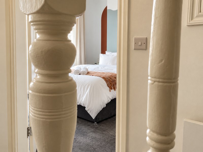 Hallway with cream-colored balusters leading to bedroom with made bed and carpet flooring