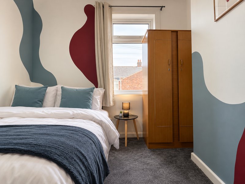 Modern bedroom with double bed, blue and red wall art, wooden wardrobe, and window view