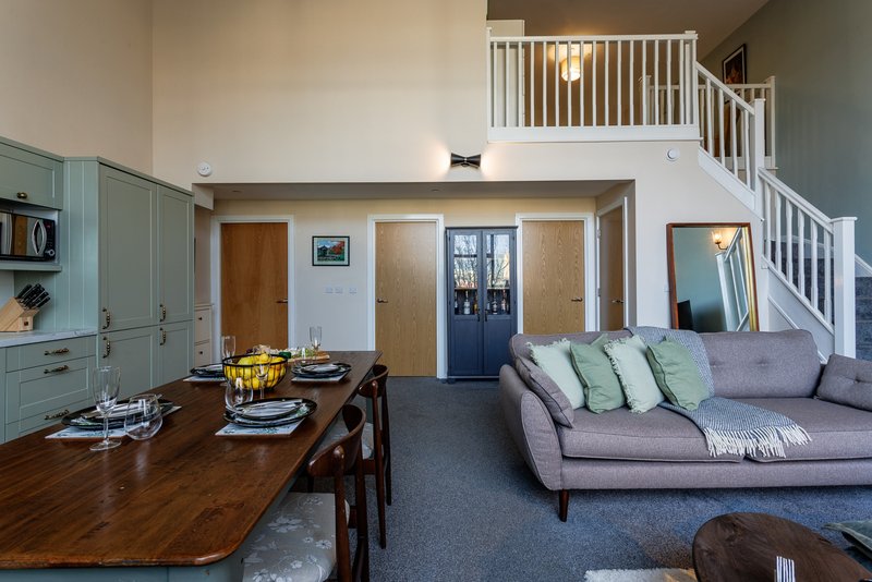 Open-plan living area with dining table, grey sofa, kitchen cabinets and staircase to mezzanine
