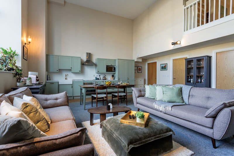 Open-plan living area with gray sofa, brown armchair, kitchen with green cabinetry and mezzanine