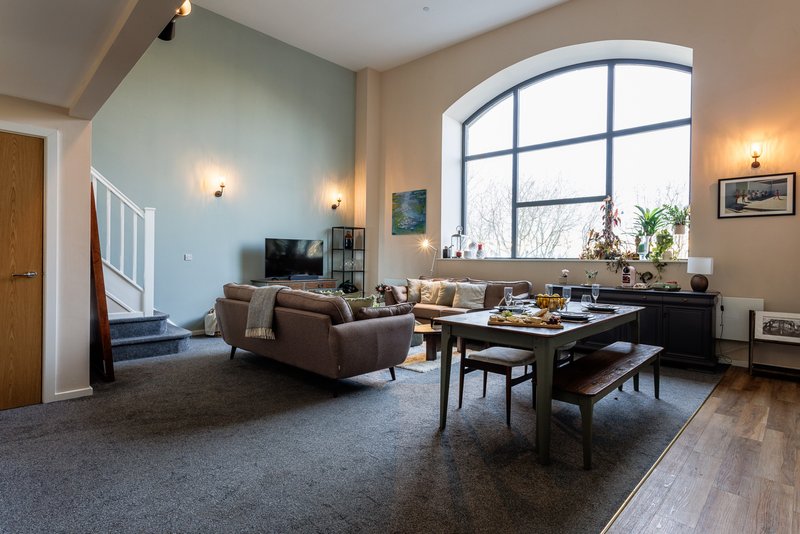 Spacious open-plan living room with large arched window, brown armchair, desk, and mixed flooring