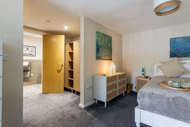 Modern bedroom with double bed, wooden dresser, artwork, and ensuite bathroom visible through doorway