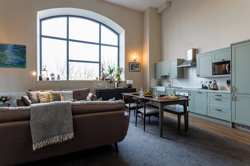 Open-plan living space with brown leather sofa, light blue kitchen units, and large arched window