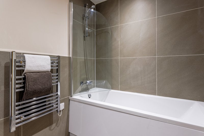 Modern bathroom with white bathtub, glass shower screen, heated towel rail and neutral tile walls
